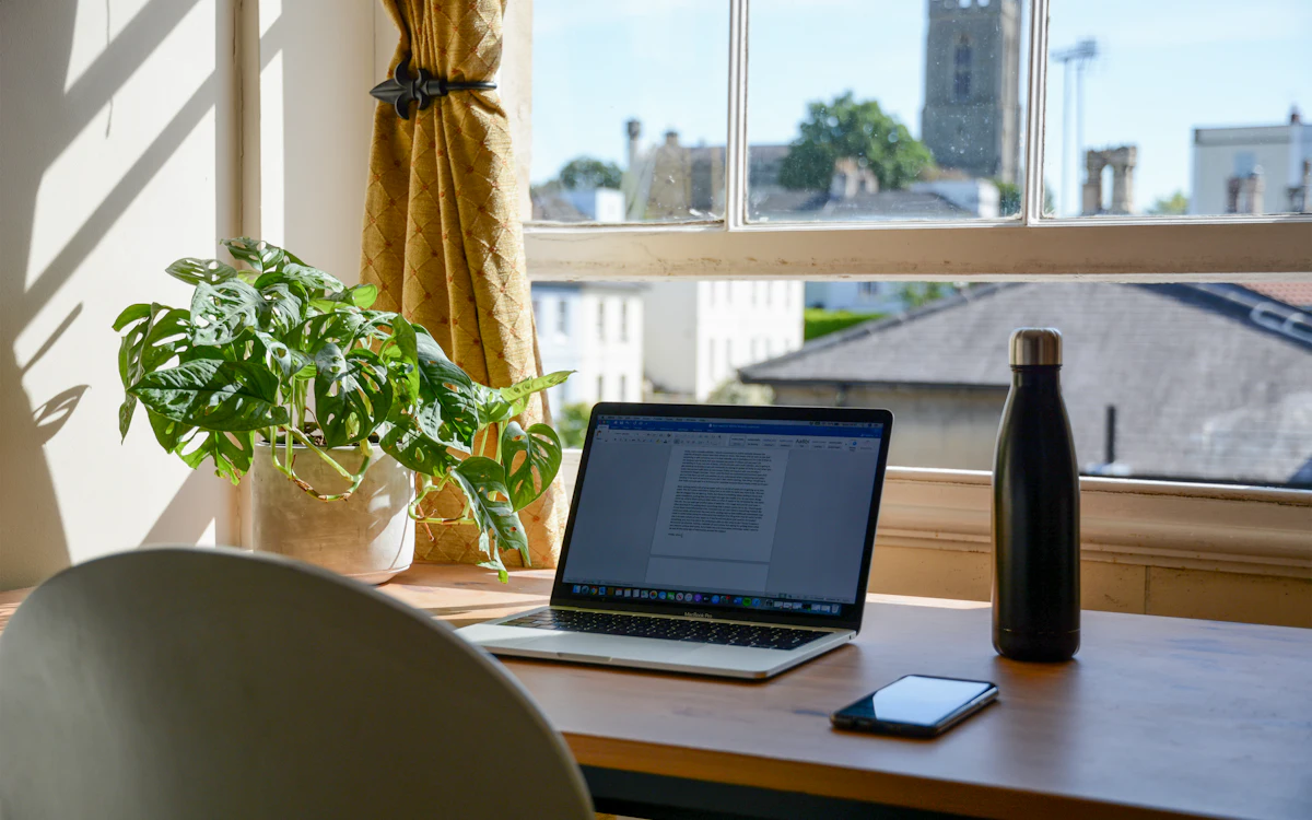 A laptop on a wooden desk by a bright window, with a plant and a water bottle