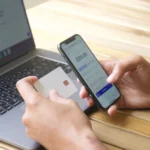 A person holding a smartphone and a bank card next to a laptop
