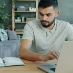 A man working on a laptop at a desk with an open notebook